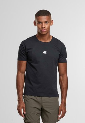 Ung man med kort hår iförd en svart åtsittande T-shirt med vitt tryck och olivgröna shorts, stående mot en enkel ljus bakgrund.