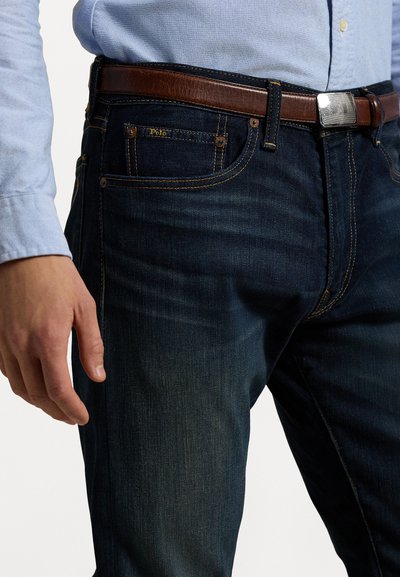 Los jeans de mezclilla azul oscuro presentan un diseño de corte recto, un sutil efecto desgastado y costuras naranjas. Se combinan con un cinturón de cuero marrón con una hebilla plateada.