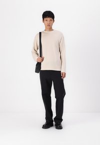 Haut beige à manches longues avec un col rond, assorti à un pantalon noir et à des chaussures noires. Un petit sac bandoulière noir est tenu sur le côté gauche.