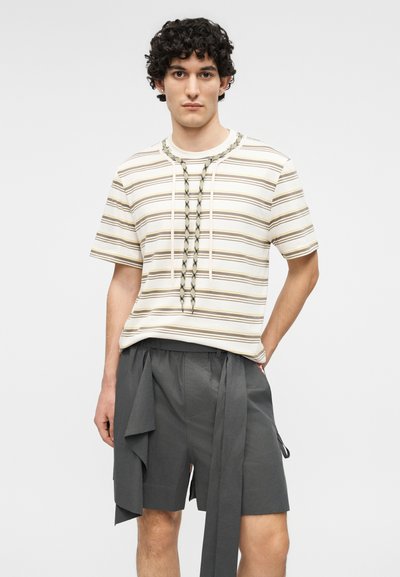 Giovane uomo con capelli ricci che indossa una camicia a maniche corte beige a righe e pantaloncini grigio scuro con dettagli di tessuto pendente.