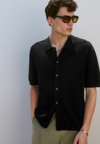Short-sleeve black button-up shirt with a textured waffle weave pattern, featuring a pointed collar and dark buttons, accessorized with sunglasses.