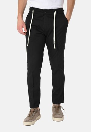 Uomo che indossa pantaloni neri slim fit con coulisse bianche e sneakers beige in suede, in piedi su uno sfondo chiaro uniforme.
