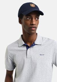Grå piké skjorte med marineblå krage, med en blå logo. Marineblå caps med et logomerke foran. Glatt stoff tekstur, klassisk passform.