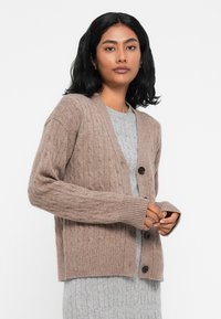 Barna, cable-knit kardigán, V nyakkivágással, bordás mandzsettákkal és gombos záródással, szürke, texturált ruha fölé viselve.