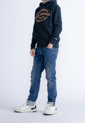 Bambino con felpa blu scuro con cappuccio e scritta, jeans blu con risvolti e sneakers bianche, in piedi con una mano in tasca contro sfondo bianco.