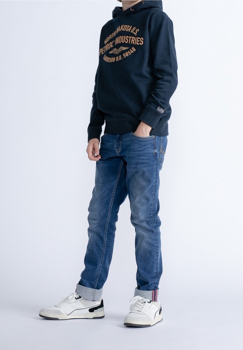 Enfant portant un sweat à capuche bleu marine avec texte, jean bleu avec revers, et baskets blanches, debout avec une main dans la poche contre un fond blanc.