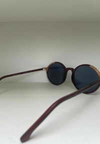 Lunettes de soleil rondes avec des verres bleu foncé, une monture bordeaux et des accents en bois sur les branches. Texture légère sur la surface de la monture.