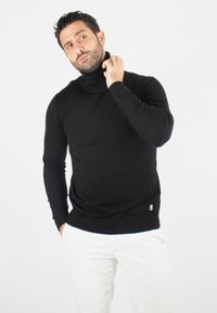 Pull à col roulé noir en maille, doté d'une coupe ajustée et de détails côtelés au col et à l'ourlet. Porté avec un pantalon blanc.