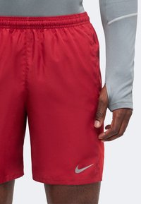 Άνδρας με κόκκινα αθλητικά σορτς Nike και γκρι μπλούζα με μακρύ μανίκι και οπή για τον αντίχειρα, στέκεται μπροστά σε ένα απλό φωτεινό φόντο.