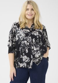 Camicetta nera con bottoni, decorata con un motivo floreale bianco e grigio, dal taglio morbido, con maniche a cuffia strutturate. Abbinata a jeans in denim scuro.