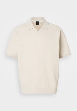 Bež polo majica s kratkimi rokavi iz pletenine, z rebrastim ovratnikom in manšetami, z diskretno našitim logotipom na prsih, prikazana na belem ozadju.