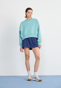 Skrojony turkusowy sweatshirt z białym logo, zestawiony z granatową spódniczką w paski. Model nosi szare trampki z jasnymi akcentami i białe skarpetki.