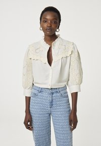 Witte blouse met kantdetails en een gerimpelde kraag, knoopsluiting aan de voorkant en uitlopende mouwen, gecombineerd met lichtblauwe gebloemde jeans.
