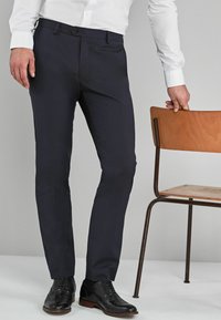 Sötétkék, testre szabott szabású nadrág sima anyagból, egyenes elejű, övbújtatókkal, fekete bőr cipőkkel és fehér inggel párosítva.