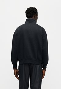 Μαύρο φούτερ με ψηλό γιακά, άνετη εφαρμογή και ριμπ μανσέτες. Υφή υφάσματος με λεία επιφάνεια, με ελάχιστες ραφές.