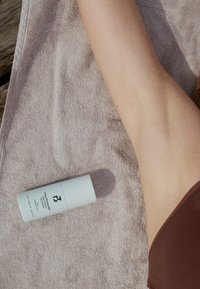 Le bras d'une personne repose sur une serviette beige à côté d'un stick de crème solaire blanc étiqueté "Pure Protection, SPF 30", sur une surface en bois à l'extérieur.