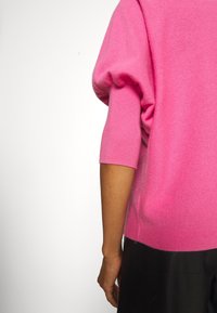 Rosa Pullover mit lässiger Passform, kurzen, puffigen Ärmeln, glatter Textur und gerippten Bündchen, kombiniert mit dunklen Hosen, von hinten betrachtet.