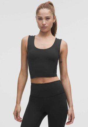 Kvinde med hestehale iført sort ærmeløs croptop og højtaljede leggings, stående foran en ensfarvet grå baggrund.