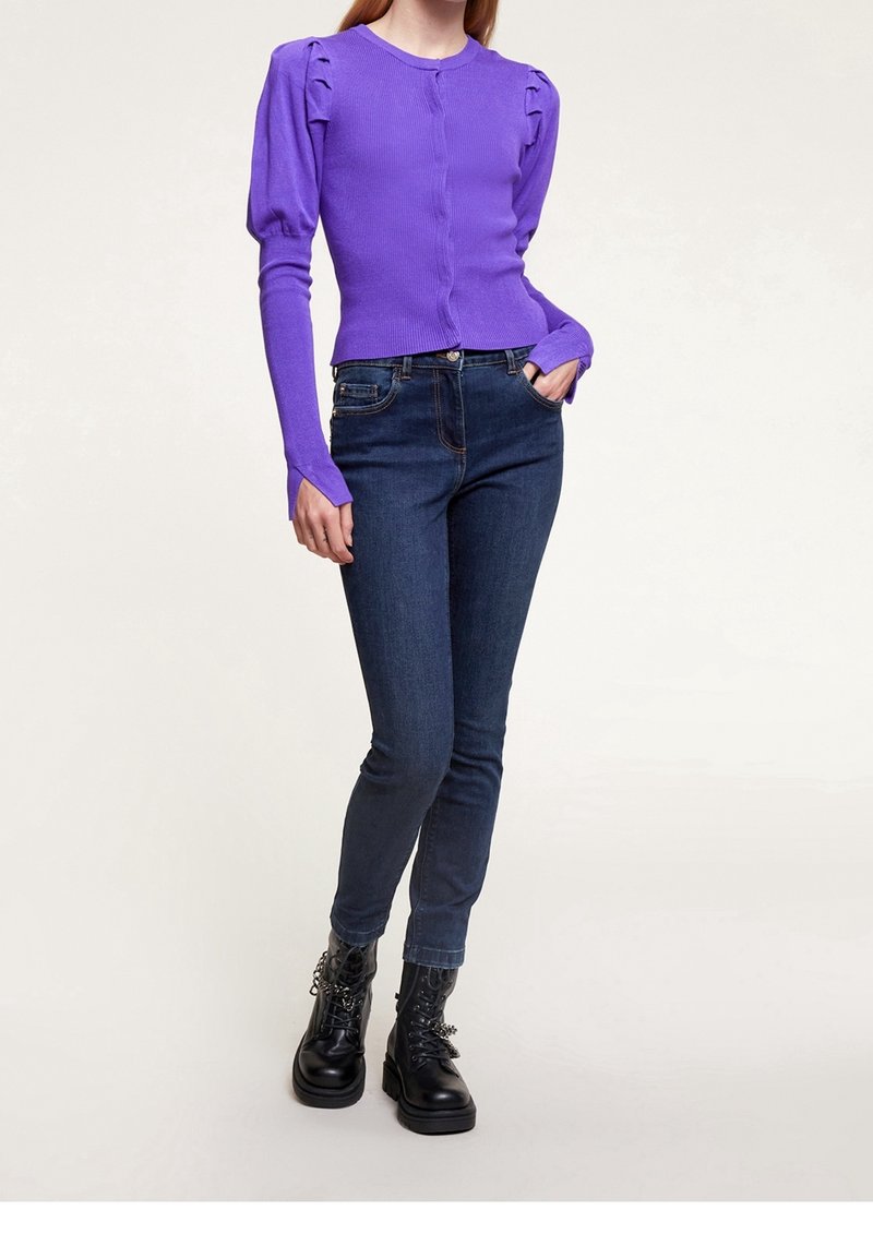 Frau trägt eine lila Cardigan mit Puffärmeln, dunkelblaue Skinny Jeans und schwarze Schnürstiefel im Combat-Stil mit Kettendetails.