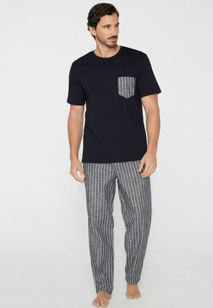 Mężczyzna w czarnej koszulce z krótkim rękawem, z kieszenią na piersi w paski, oraz w pasujących spodniach w paski, stojący boso na białym tle.