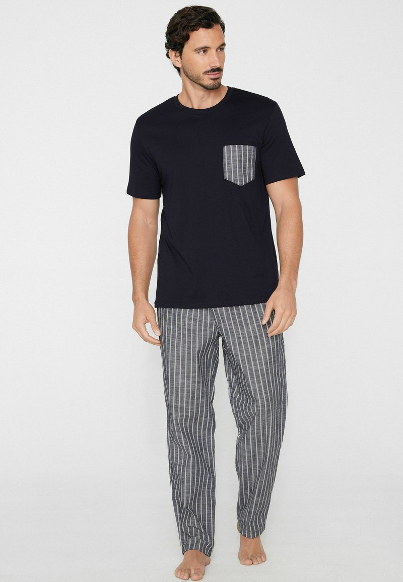 Mann trägt schwarzes kurzärmeliges Hemd mit gestreifter Brusttasche und passenden gestreiften Hosen, steht barfuß vor weißem Hintergrund.