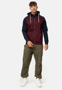 Sudadera con cremallera en color burdeos y marino con cordones, que presenta un diseño ajustado y puños acanalados. Combinada con pantalones cargo verdes y zapatillas blancas.