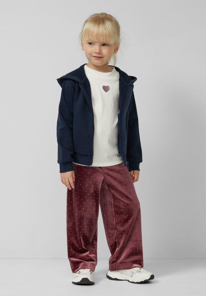 Sudadera con cremallera en color navy, camiseta blanca con gráfico de corazón, pantalones de terciopelo burdeos con puntos blancos, zapatillas deportivas blancas.