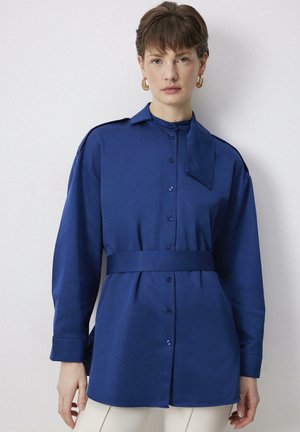 Nő hosszú ujjú királykék gombos blúzban, derékövvel és arany karika fülbevalóval, egyszerű fehér háttér előtt állva.