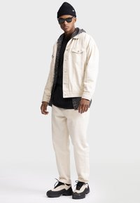 Cream denim jacket with gray hoodie lining, black t-shirt, cream joggers, and black and beige sneakers, accompanied by black sunglasses and beanie.