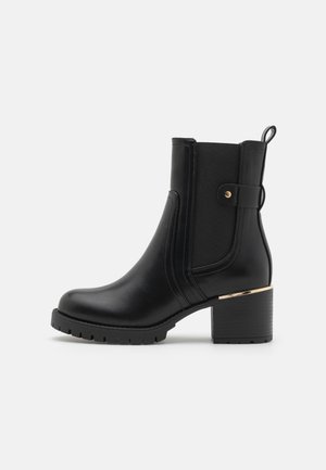 Botte noire à la cheville en cuir lisse avec un panneau texturé, un talon bloc et des accents dorés. Équipée d'une bride pour un enfilage facile.