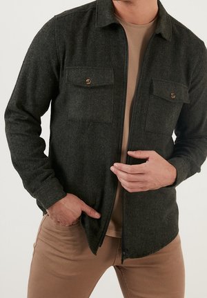 Mann trägt dunkelgraue, geknöpfte Hemdjacke über einem beigen strukturierten Hemd und hellbraunen Hosen, Hand in der Tasche, richtet die Vorderseite der Jacke.