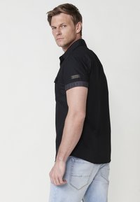 Camisa negra de manga corta con puños enrollados, que presenta un bolsillo en el pecho. Tejido suave y un diseño ajustado, combinada con jeans de lavado claro.