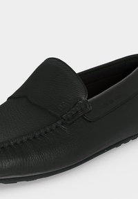 Μαύρο δερμάτινο loafer χωρίς κορδόνια με υφή στην άνω επιφάνεια, ραφές μοκασίνου κατά μήκος της μύτης και σόλα χαμηλού προφίλ.