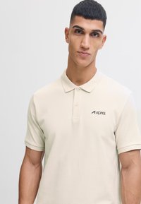Beige polo shirt met een textuurpatroon, met een drieknoopsluiting en logo op de linkerborst. Korte mouwen en casual fit.