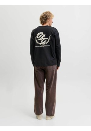 Personne blonde portant un t-shirt noir à manches longues avec un texte graphique blanc dans le dos, un pantalon marron ample et des chaussures claires, debout de dos.