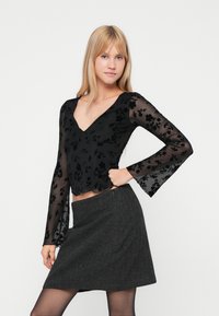 Blusa preta transparente com bordados florais, com decote em V e mangas largas, emparelhada com uma saia em linha A cinza com padrão de espinha de peixe.