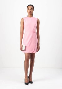 Robe sans manches rose avec une coupe ajustée, longueur genou, et détail de nœud sur le côté. Associée à une pochette texturée blanche et des talons noirs.