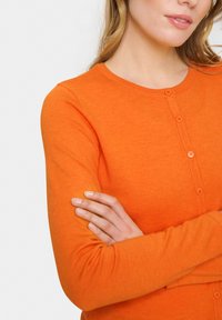 Ženska v svetlo oranžni dolg rokav z gumbi, s prekrižanimi rokami, vidna del njenega obraza in zgornjega dela telesa na beli podlagi.