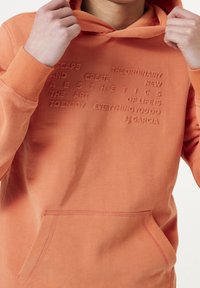 Eine Person hält die Kapuze eines orangefarbenen Sweatshirts, auf dem in erhobener Schrift steht: "Entfliehen und Ästhetik schaffen, die Kunst des Lebens besteht darin, alles, was du tust, zu genießen."