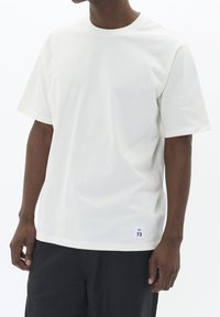 Weißes Baumwoll-T-Shirt mit klassischem Rundhalsausschnitt, kurzen Ärmeln und einem kleinen Logetikett am Saum. Glatte Textur und lockere Passform.