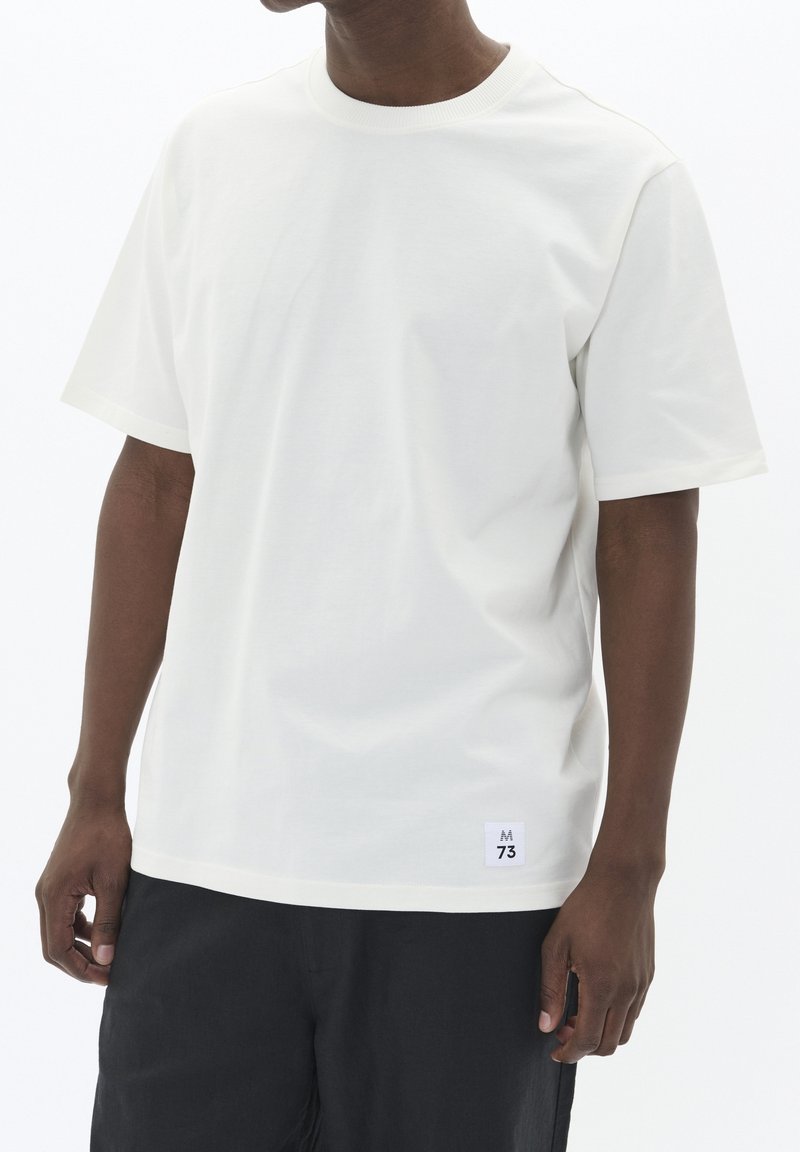 Weißes Baumwoll-T-Shirt mit klassischem Rundhalsausschnitt, kurzen Ärmeln und einem kleinen Logetikett am Saum. Glatte Textur und lockere Passform.