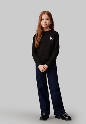 Camicia nera di cotone a maniche lunghe con logo sul petto, abbinata a pantaloni in denim scuri a gamba larga e scarpe nere. Design semplice, vestibilità classica.