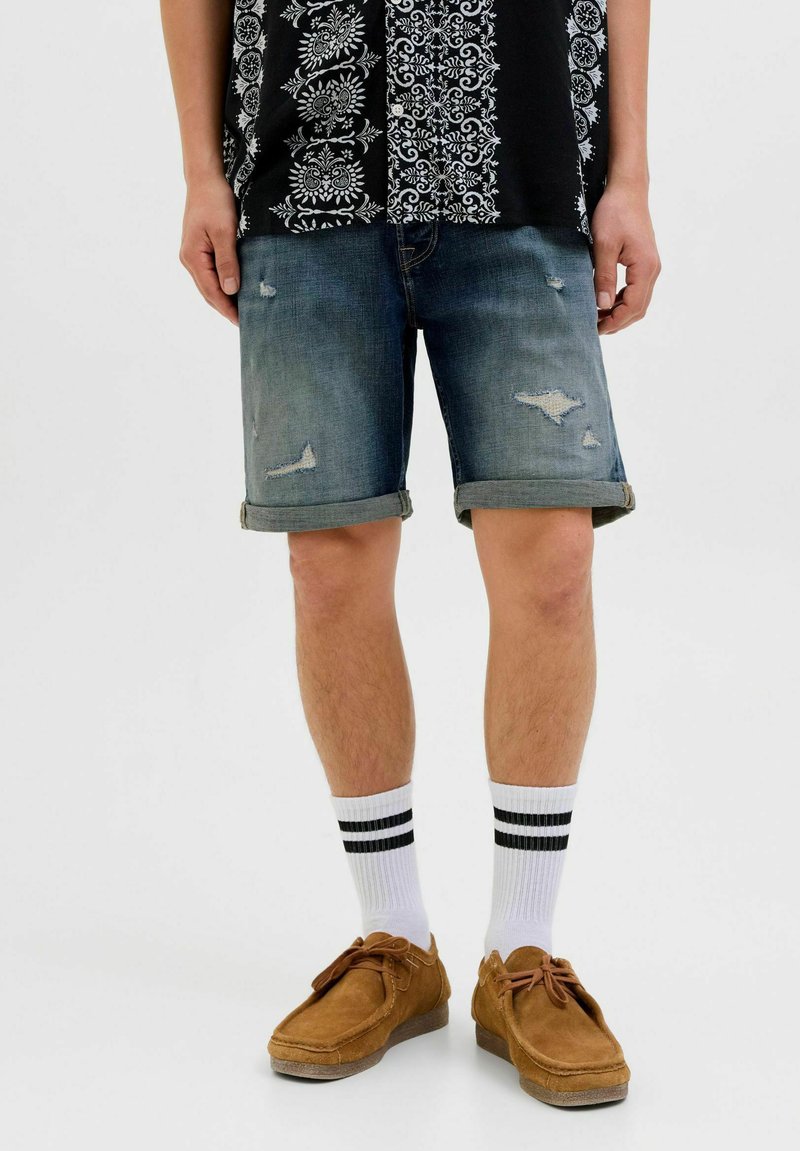 Shorts en denim avec un design déchiré, délavage bleu clair et ourlets retroussés, associés à des chaussettes blanches avec des rayures noires et des chaussures marron.