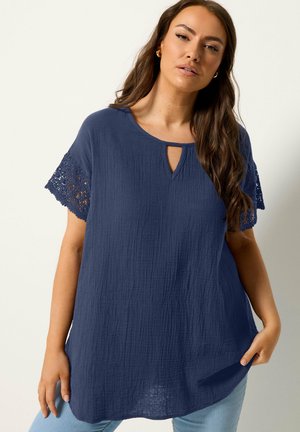 Donna con capelli castani lunghi che indossa una camicetta blu navy strutturata con maniche all'uncinetto e jeans azzurri, posa davanti a uno sfondo chiaro.