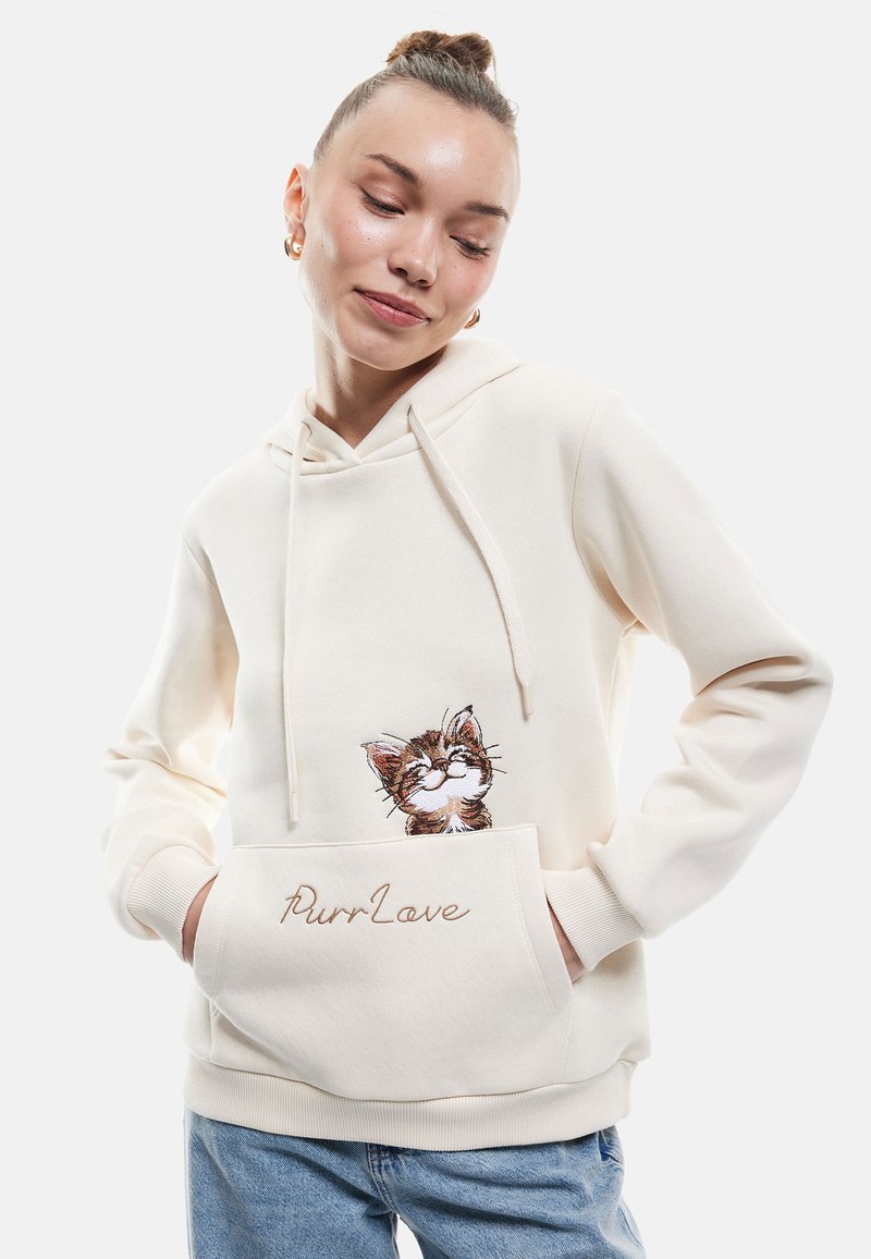 Sudadera crema con un bolsillo frontal, que presenta un diseño de gato bordado y el texto "PurrLove", hecha de una tela suave con un corte relajado.