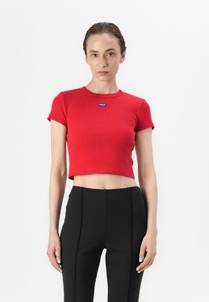 Red cropped t-shirt made of ribbed fabric featuring a round neckline and a blue label at the chest. Paired with high-waisted black pants.
