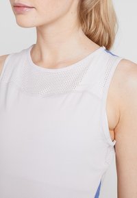 Débardeur de sport gris clair avec des détails en mesh aux épaules, présentant une texture douce et des accents bleus sur les panneaux latéraux.