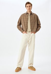 Camisa de cuadros marrón sobre una camiseta beige, combinada con pantalones holgados color crema y zapatos mocasín marrón claro.