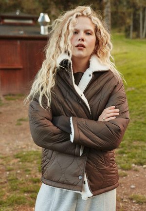 Giacca marrone trapuntata con colletto in shearling bianco, caratterizzata da bottoni a scatto e una vestibilità rilassata, indossata sopra un top scuro e pantaloni sportivi grigi.
