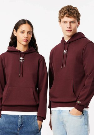 Sudaderas burdeos con bolsillos frontales y cordones; con un pequeño logo de Lacoste en el pecho y puños de canalé. Lleva puestos unos jeans de mezclilla claros.